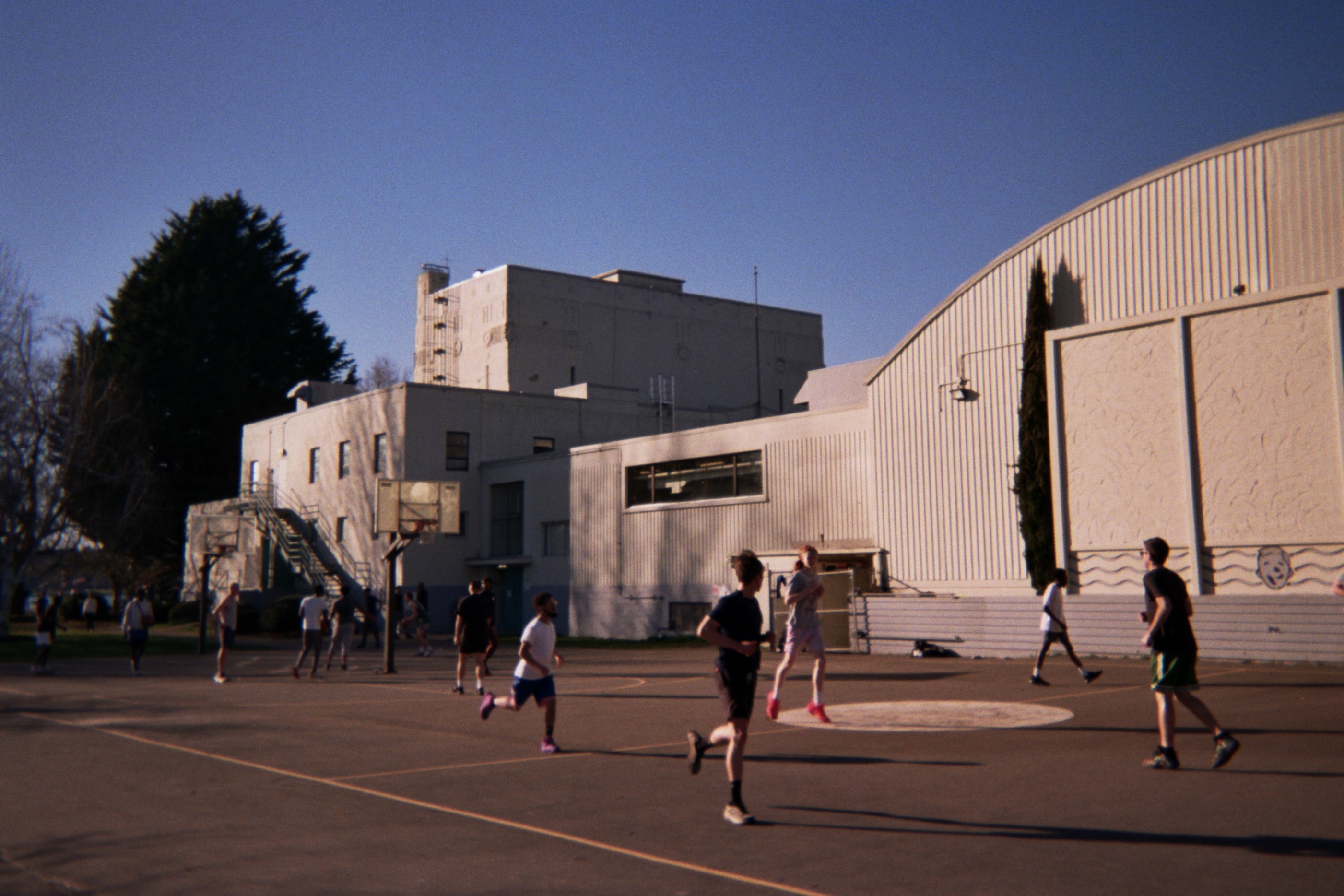 People playing basketball on an outdoor court next to a large institutional building with white walls and a curved roof.